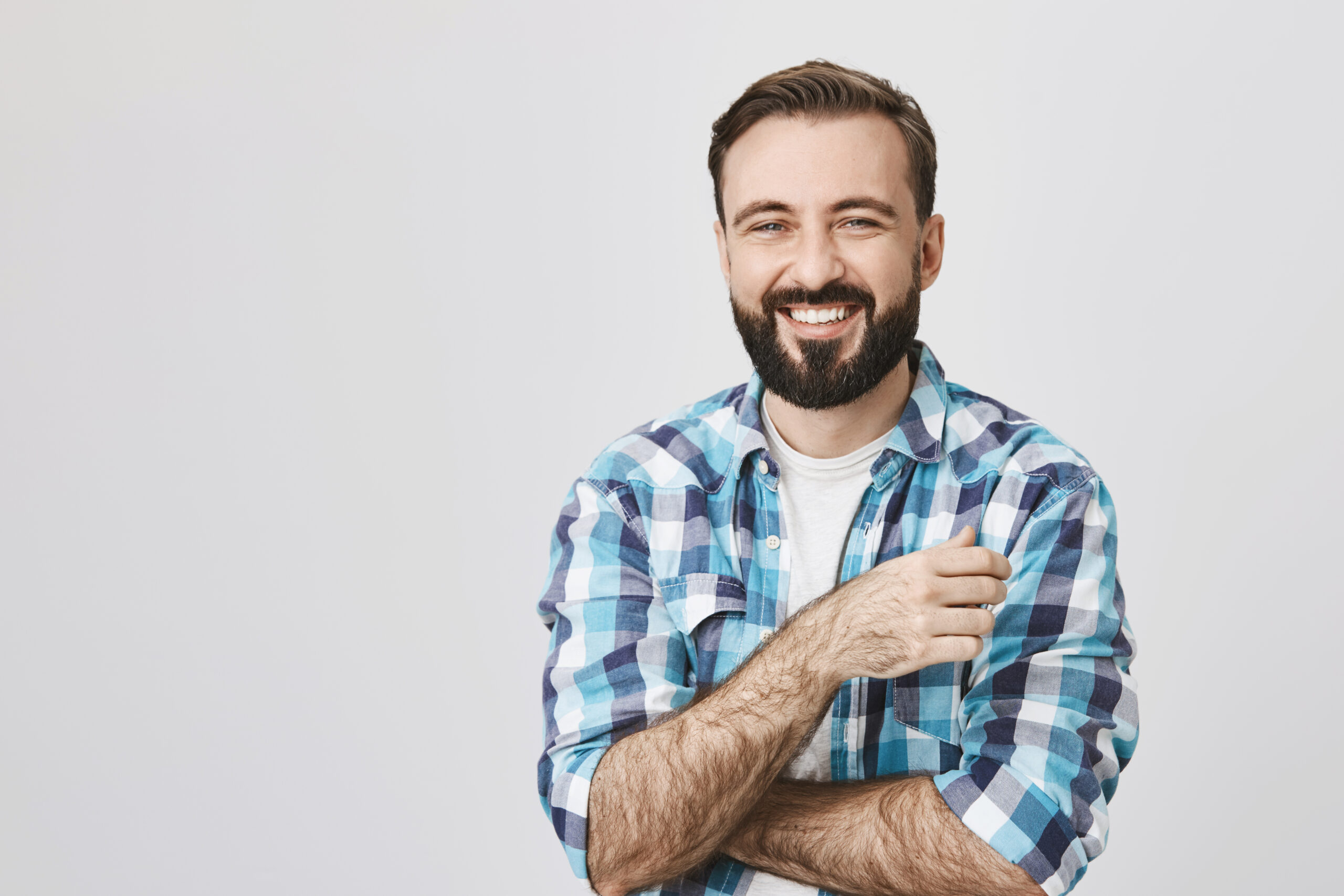 Portrait of adult bearded man with shiny smile and crossed hands, looking at camera, over gray background. Guy laughs nervously in front of father-in-law, meeting him for the first time.